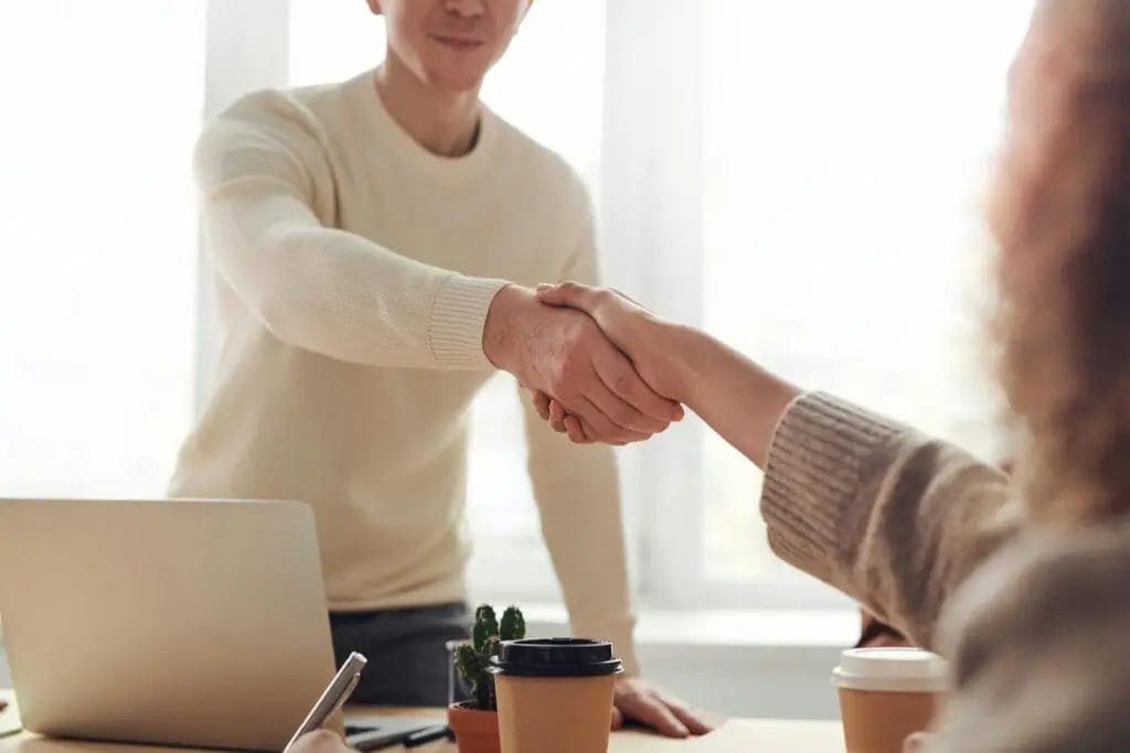 Two people are shown shaking hands indoors, suggesting a successful meeting or agreement. One person is seated at a table with coffee cups, a small plant, and a laptop in the background. Both are dressed in light-colored sweaters, bridging the gap to enhance customer experience.