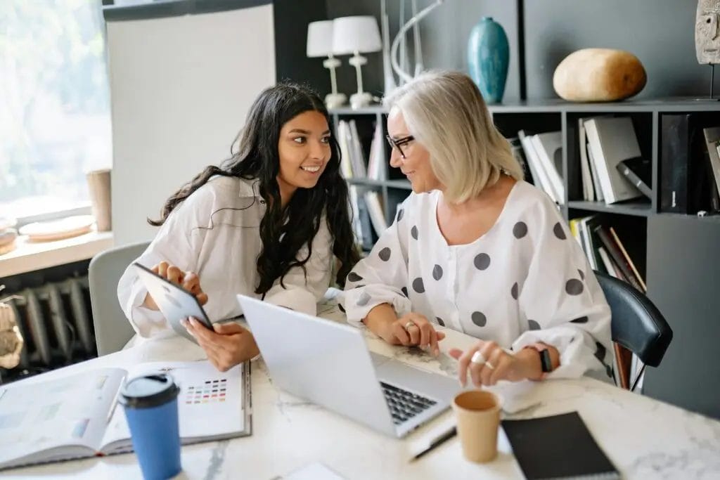 Two women are sitting at a table with a laptop and various notebooks. One woman with dark hair holds a tablet, while the other with gray hair looks at her, smiling. They appear to be engaged in a conversation about User Experience (UX) Design. Coffee cups sit on the table.