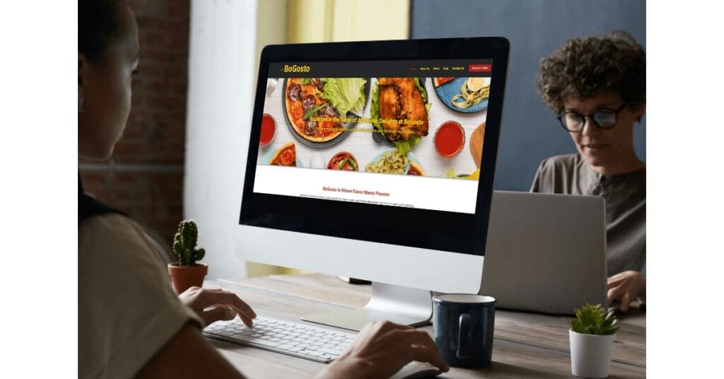 Two people working at desks with laptops. The person in the foreground has a desktop displaying a website for BoGusto, featuring various dishes and a menu. Small plants and cups are on the desks. The background is partially blurred with a dark blue wall.