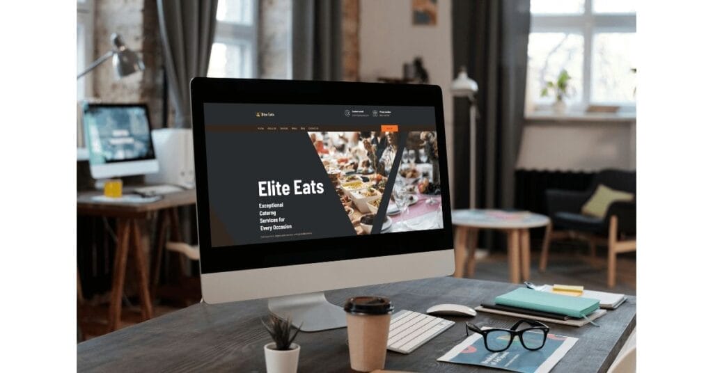 An office desk setup featuring a desktop computer displaying the homepage of "Elite Eats," a catering service. The desk also has a smartphone, a coffee cup, and various documents. In the background, there are windows and another desk with a laptop in a cozy office space.