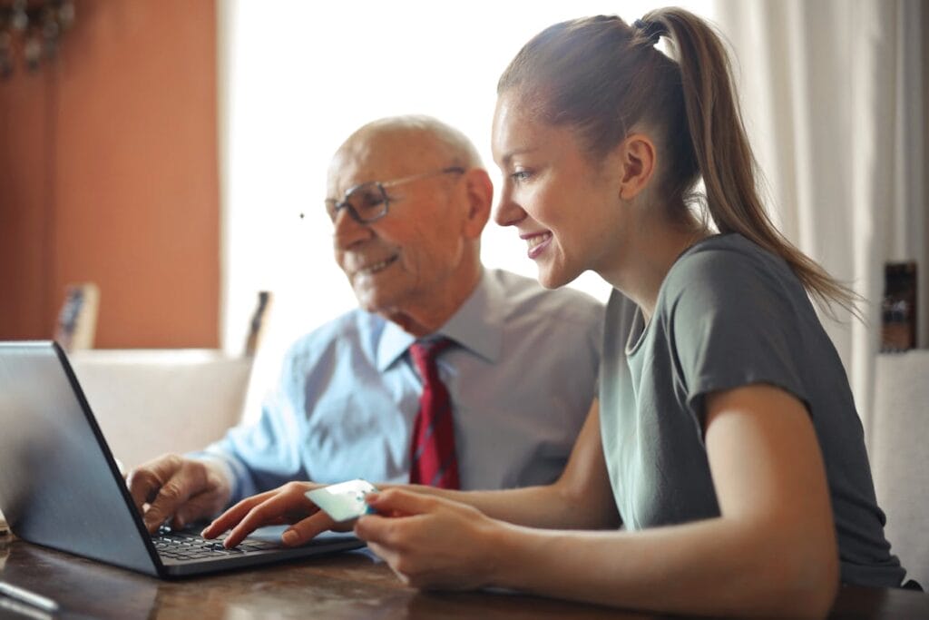 A young woman with a ponytail sits at a table with an elderly man wearing glasses and a tie. Both are smiling and looking at a laptop screen, where they explore digital marketing hacks. The young woman is holding a credit card in her right hand. Sunlight filters through a window in the background.