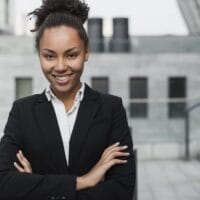 A confident person with curly hair in a bun is smiling while standing outdoors. They are wearing a black suit and white shirt, with arms crossed. The background features a modern building with glass and concrete elements and a staircase. A confident person with curly hair in a bun is smiling while standing outdoors. They are wearing a black suit and white shirt, with arms crossed. The background features a modern building with glass and concrete elements and a staircase.
