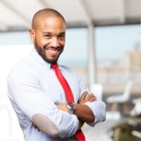 A man with a beard and closely shaved head smiles confidently, wearing a light blue dress shirt with dark patches on the elbows, a bright red tie, and a silver watch. He stands in an office space with modern furniture and a large window in the background. A man with a beard and closely shaved head smiles confidently, wearing a light blue dress shirt with dark patches on the elbows, a bright red tie, and a silver watch. He stands in an office space with modern furniture and a large window in the background.